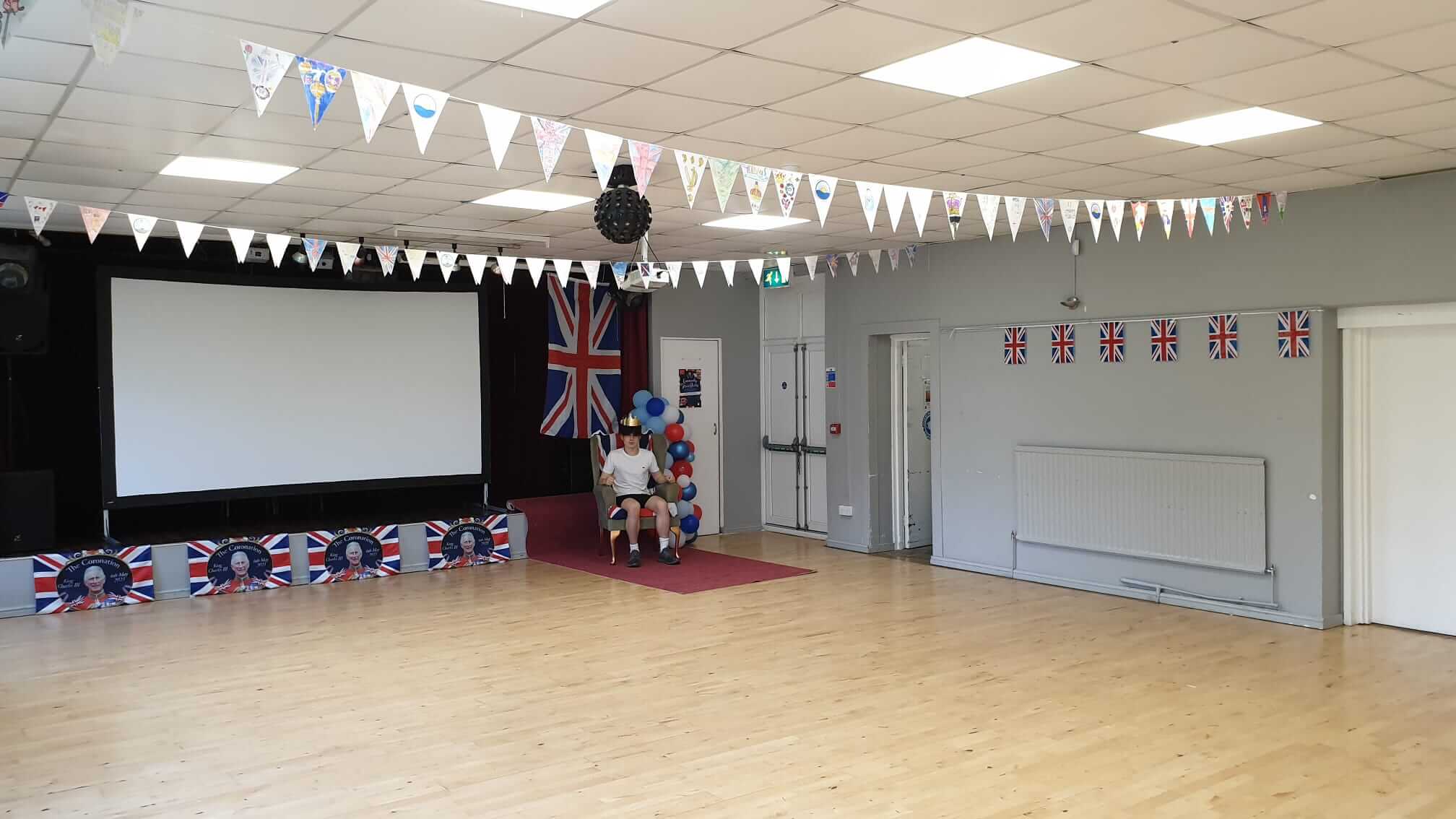 a community hall decorated for the coronation with union jacks. In the corner of the room there is a throne