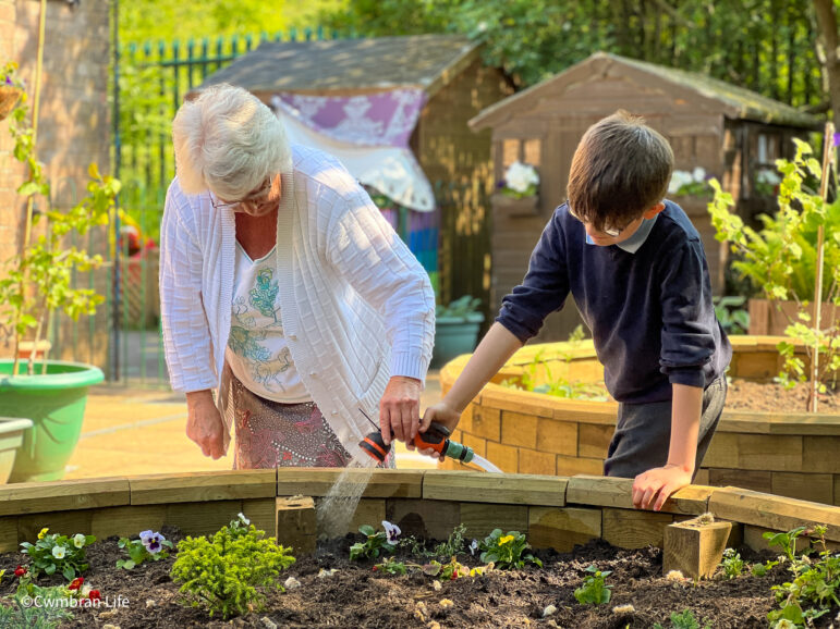 a boy waters plants watched by an older woman