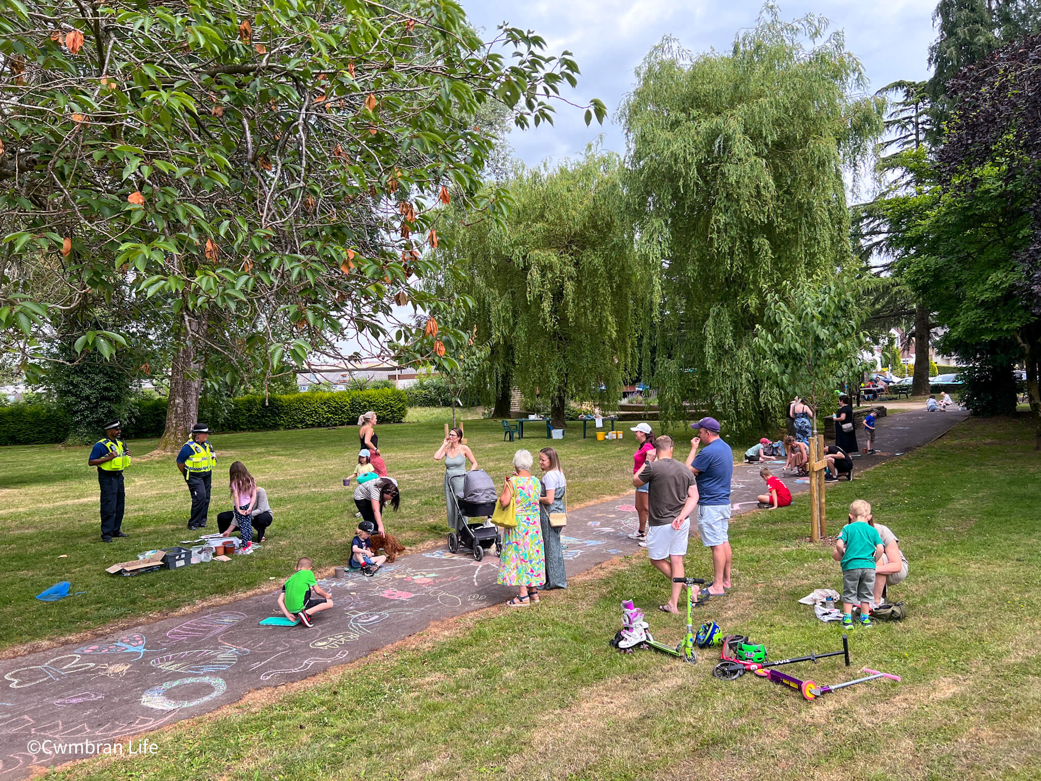a group of adult and children stand in park while some draw in chalk on a path