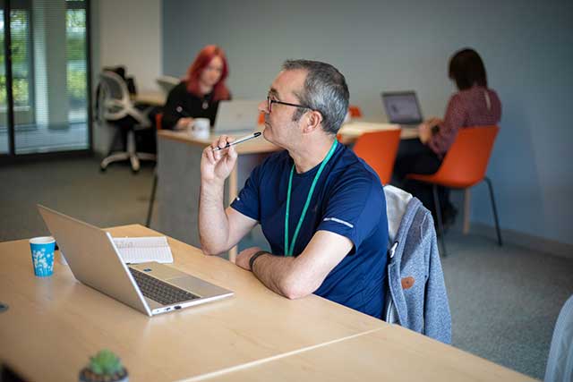 a man working on a laptop at a desk- two people work at desk behind him