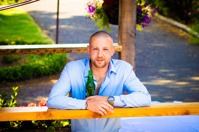 a man leaning on wooden post looking at camera while holding a beer