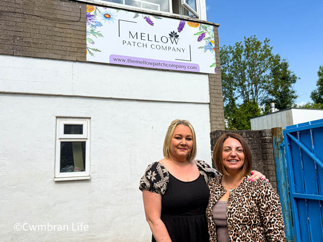 Two women outside an office. A sign on the wall says The Mellow Patch Company