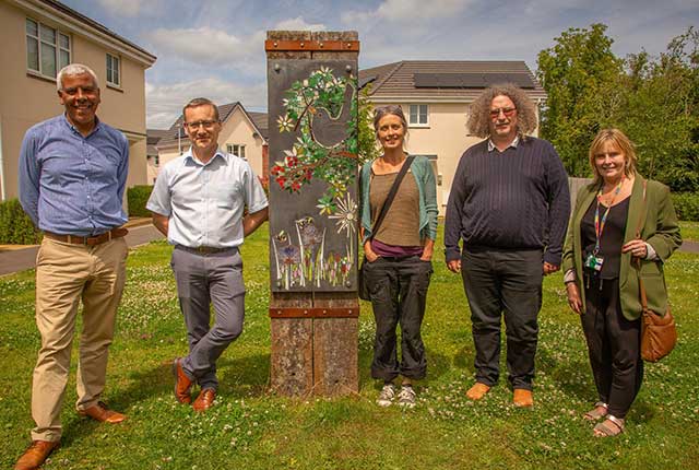 three men and two women stood by a mosaic mural showing a bird holding twigs and a river