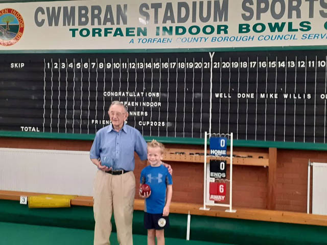 a 98 year old man stood by an 8 year old boy on an indoor bowling green