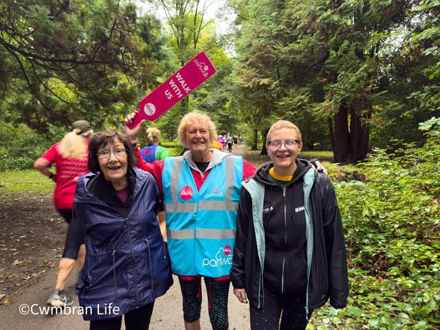 three women smile during a parkrun. One holds a sign saying 'walk with us'