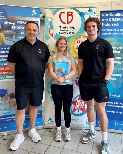 Two men and a woman stood by a board advertising the CB training services swim school