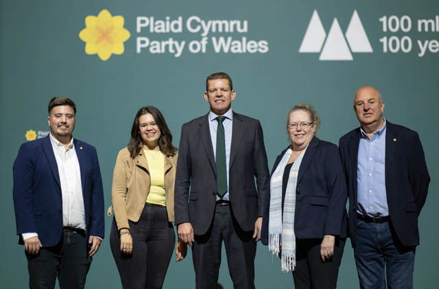 three men and two women in front on a plaid cymru sign