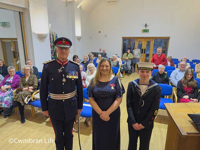 a woman stood inbetween a bridgadier and a sea cadet with rows of people sat behind them