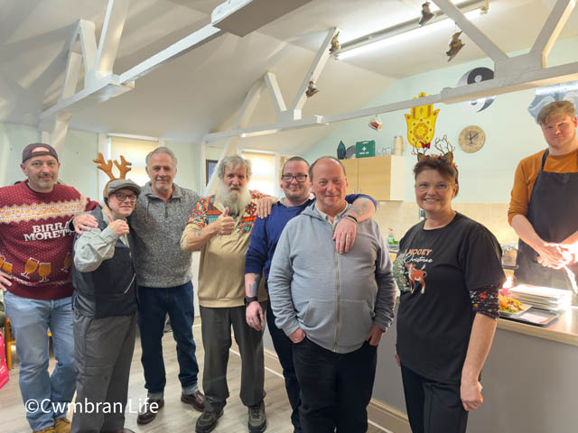 Eight people in a kitchen, some wearing Christmas jumper and hats