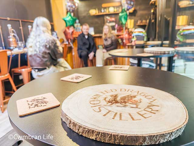 a table in a bar with a mat saying 'kilted dragon distillery'- in the background, blurred are a couple sat on chairs by the bar in front of a woman