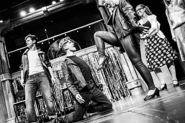 a woman puts her foot against a kneeling man's chest in a scene on stage from Grease the musical