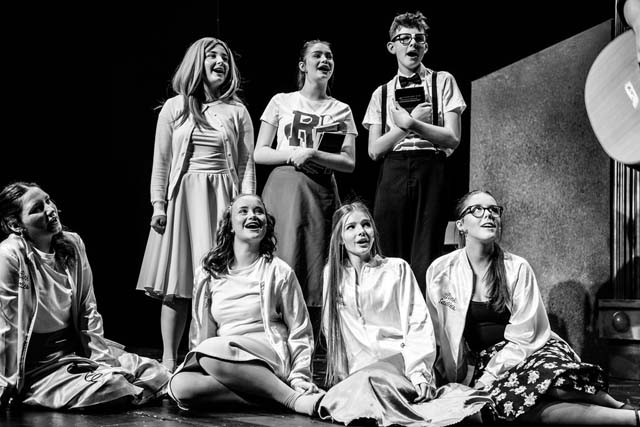 a group of teenagers sat on stage in a scene from grease the musical