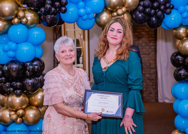 two women stood in front of blue and black balloon arch holding a certificate