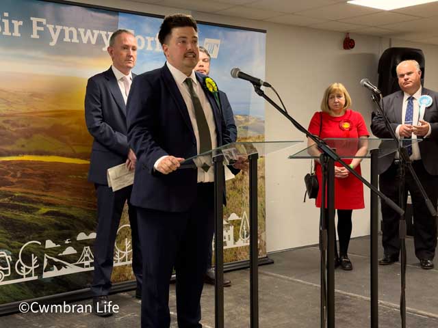 a man gives a speech at a lecturn with four people in the background