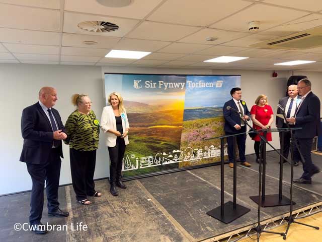 seven people on a stage with a sign saying 'Sir Fynwy Torfaen' behind them