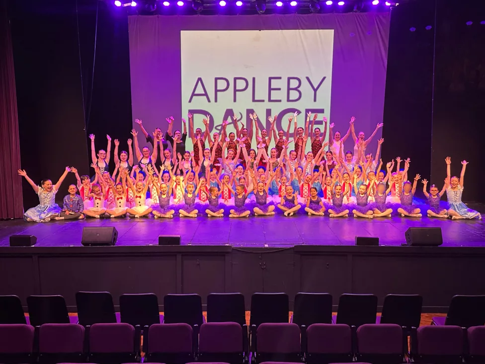 a cast of a ballet school sat as a team on a theatre stage