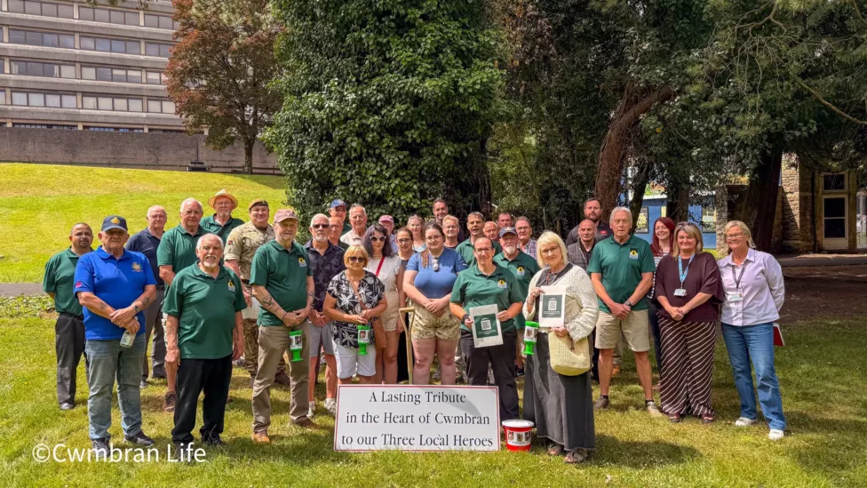 a group of people stood on a grassed area next to card saying 'lasting tribute in the heart of cwmbran to our local heroes'