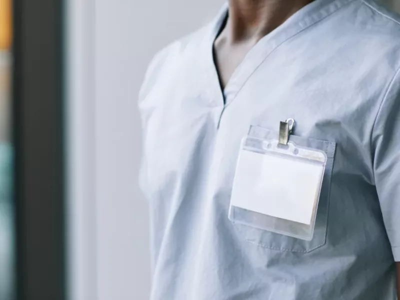 A medical professional is depicted from the chest up, wearing light blue scrubs. A clear plastic ID badge is clipped to the scrub top.
