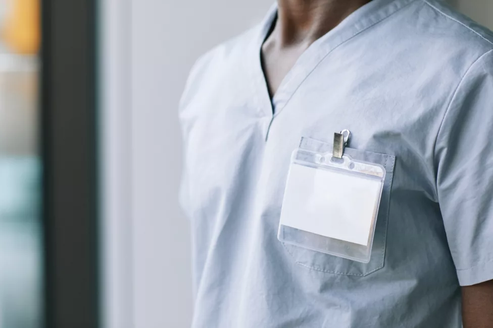A medical professional is depicted from the chest up, wearing light blue scrubs. A clear plastic ID badge is clipped to the scrub top.